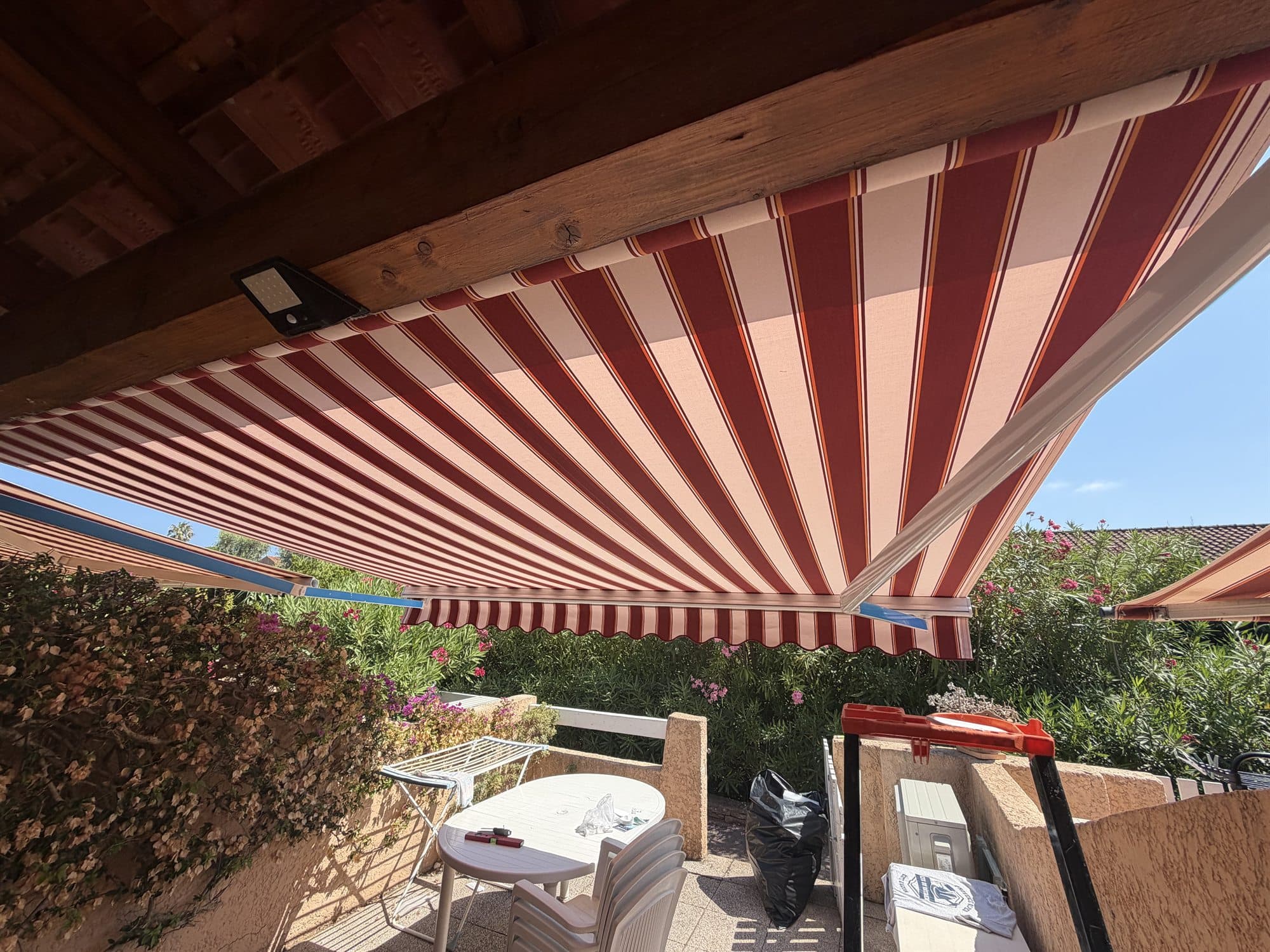 Banne à rayures rouges et blanches fixée sous des poutres en bois sur une terrasse fleurie